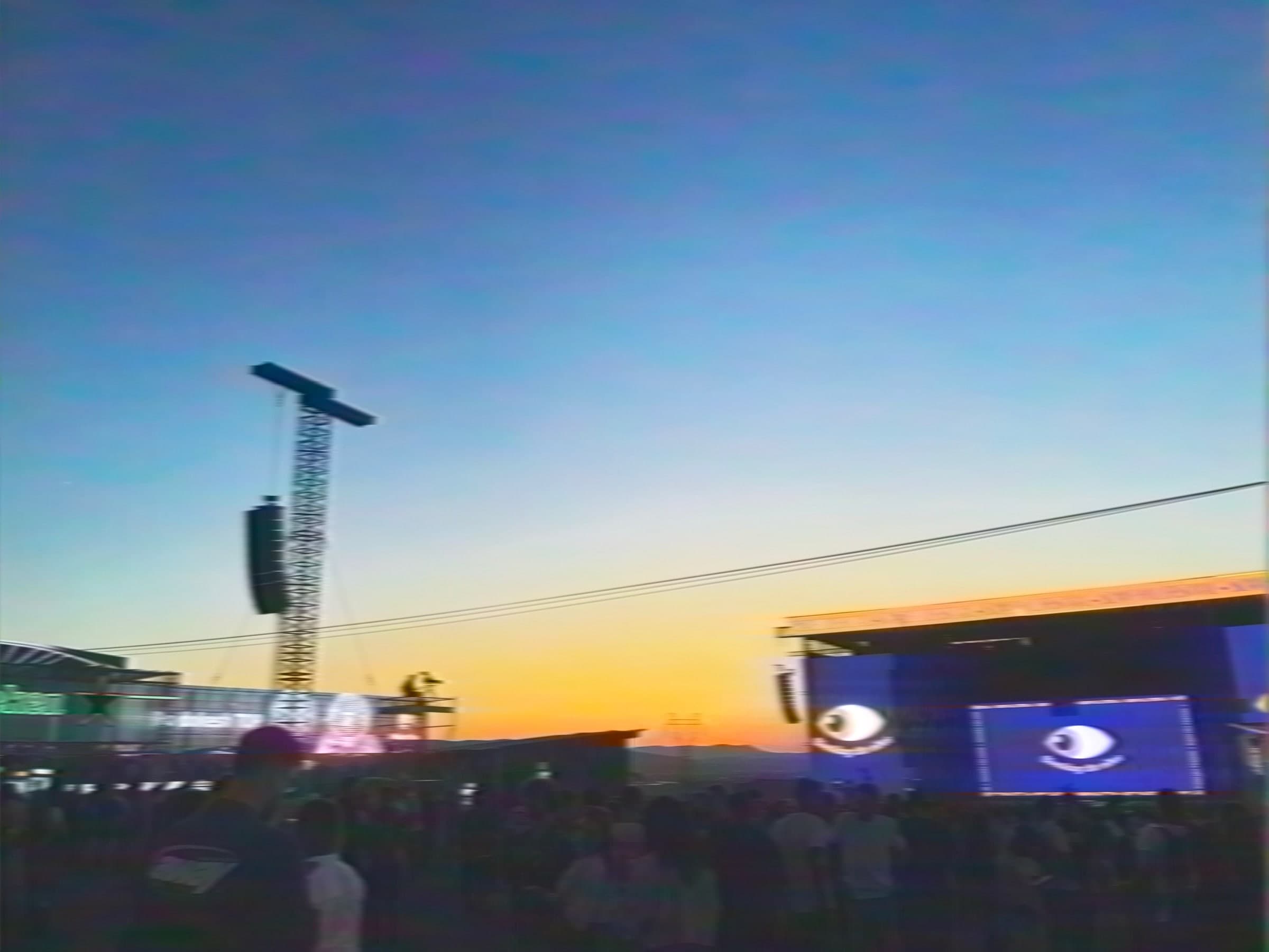 Açık hava festivali: kalabalığın üstünde asılı hoparlörler, dev göz görselli LED ekran ve turuncudan maviye giden gökyüzü.