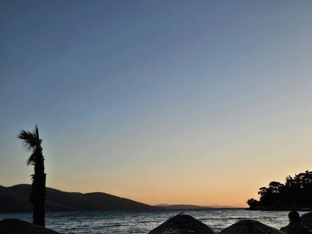 Gün batımında deniz, palmiye ağacı silueti ve dağların arkasından süzülen altın sarısı ışık.