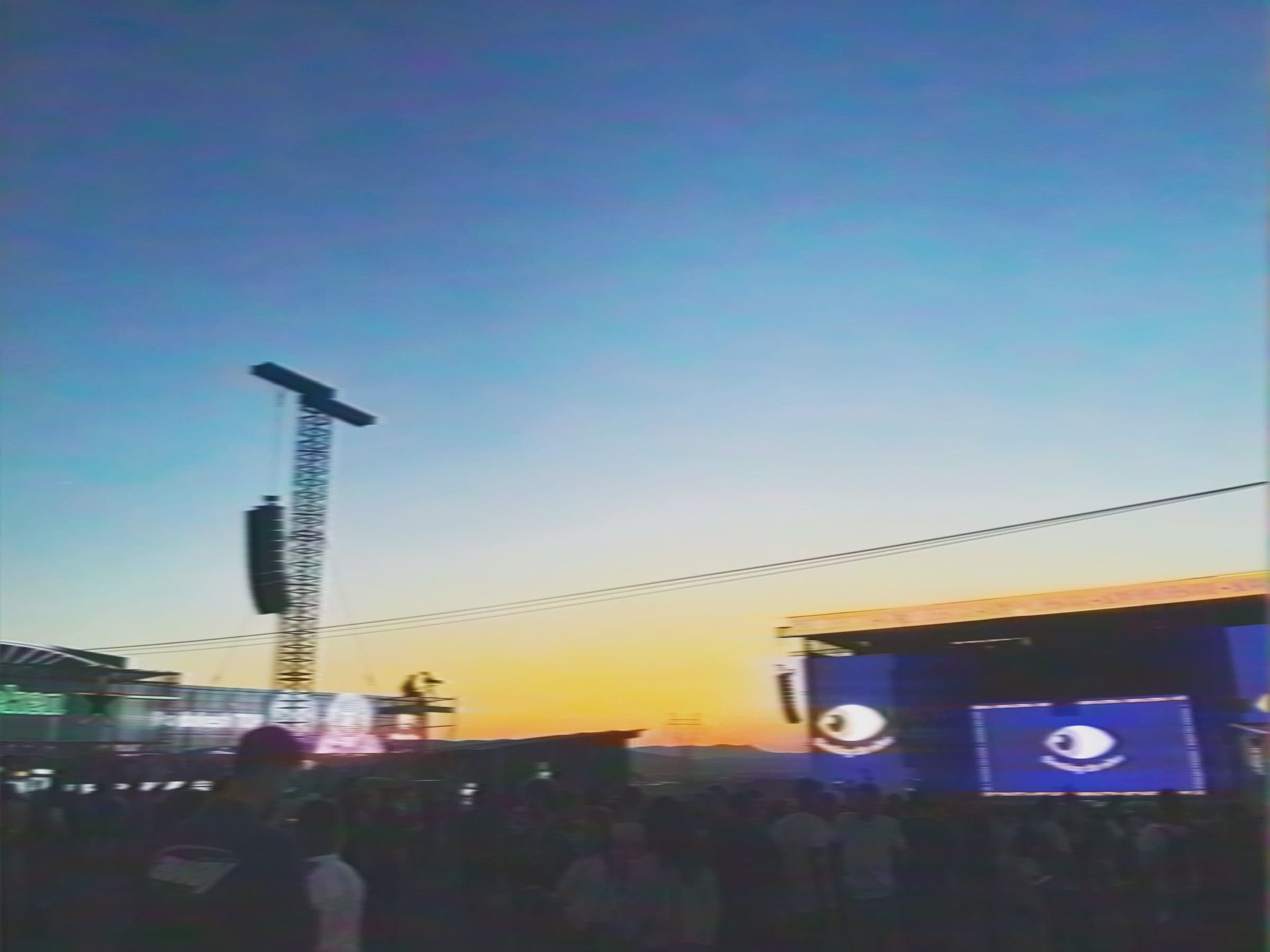 Açık hava festivali: kalabalığın üstünde asılı hoparlörler, dev göz görselli LED ekran ve turuncudan maviye giden gökyüzü.